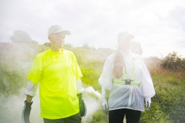 FI: Kaksi ihmistä seisoo avarassa maisemassa aurinkoisena päivänä. Taustalla ja ihmisten kasvojen edessä on savua. EN: Two people stand in an open landscape on a sunny day. There is smoke in the background and in front of the people's faces. Outo Olo
